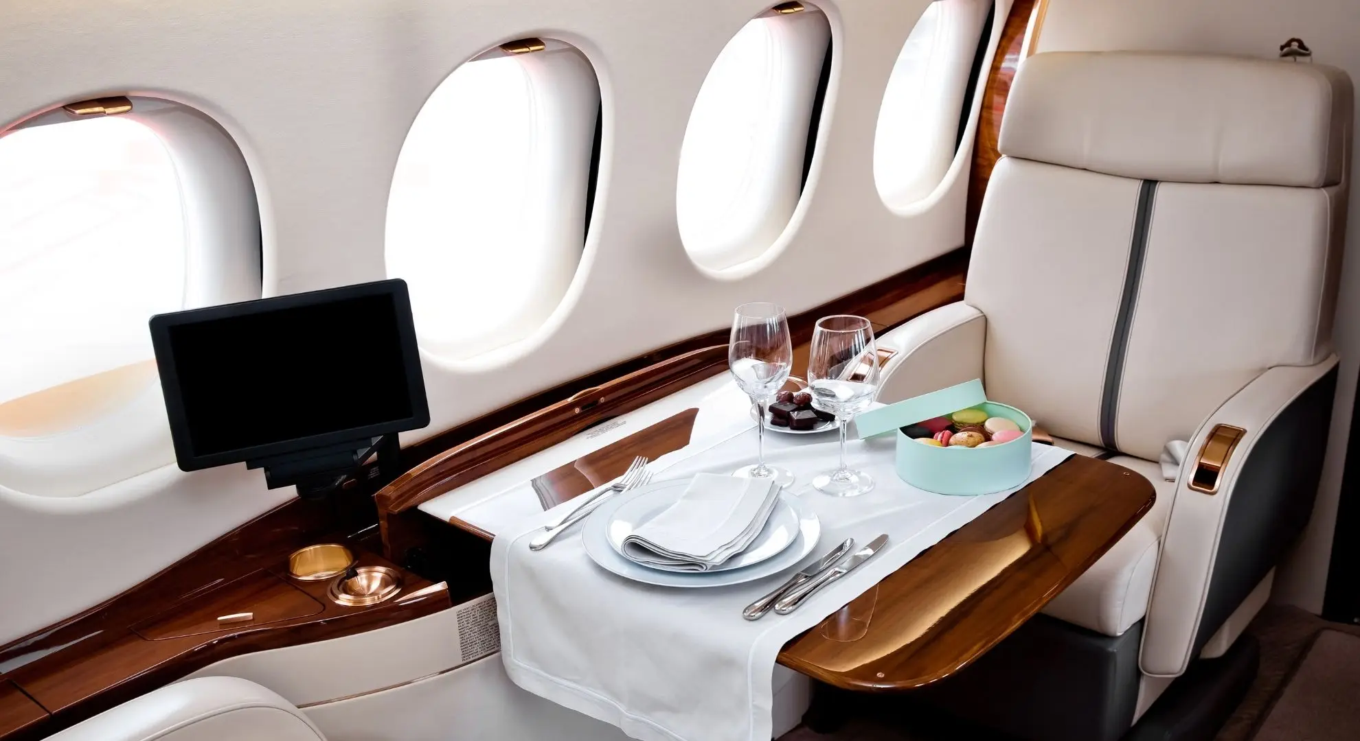 Private jet cabin dining setup with fine china, wine glasses and a fold-out table beside cream leather seats and oval windows