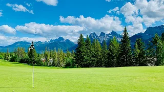 Pristine golf course with manicured green fairway and flag, surrounded by dense pine forests and dramatic mountain peaks under blue sky