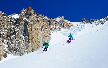 Two skiers descending pristine powder snow slopes beneath dramatic rocky peaks at Jackson Hole