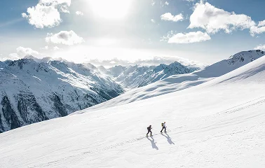 Two skiers descending pristine powder slopes with snow-capped mountain peaks under bright alpine sun