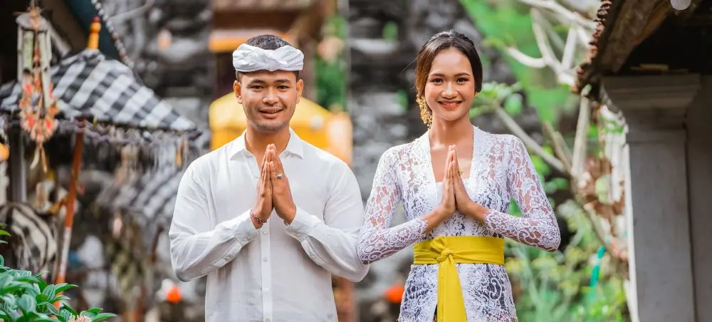 Balinese couple in traditional white ceremonial attire performing namaste greeting at ornate temple