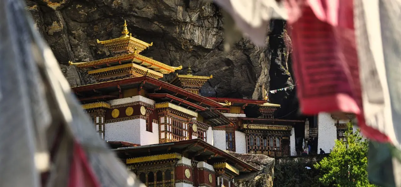 Traditional Bhutanese monastery with golden roofs nestled against dramatic mountain cliffs, viewed through colorful prayer flags