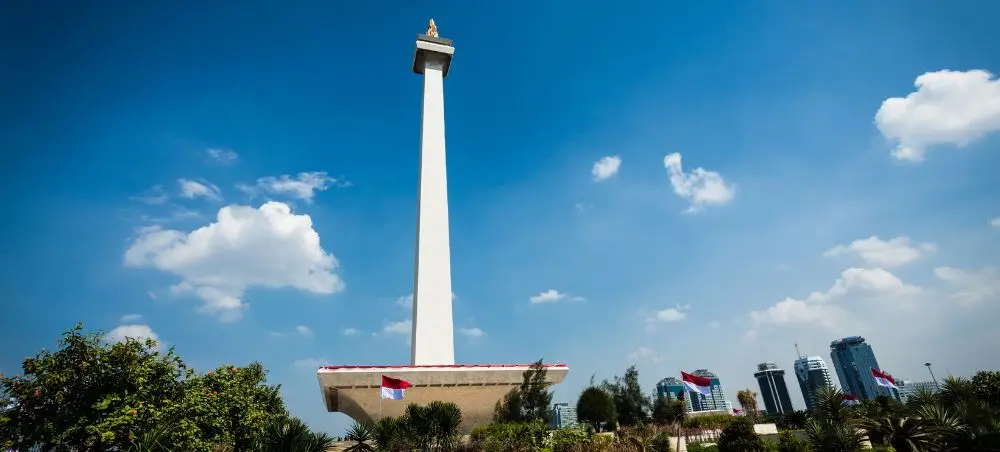 National Monument towering above Jakarta's skyline with modern buildings and lush tropical gardens below