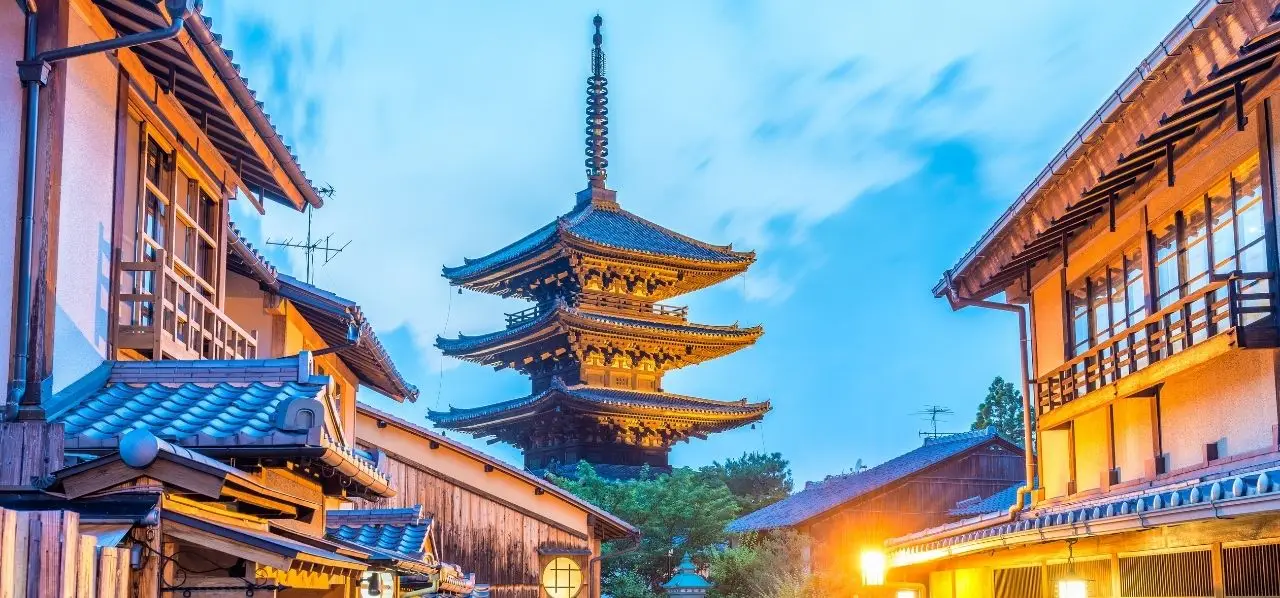 Historic Kyoto pagoda illuminated at dusk surrounded by traditional wooden buildings and blue tiled roofs