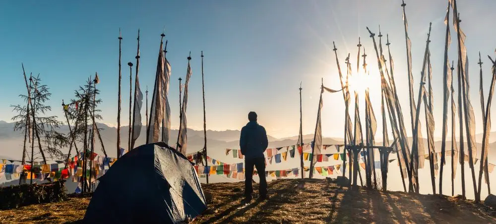 Traveler camping among colorful prayer flags at sunrise in Paro, Bhutan with mountain peaks backdrop
