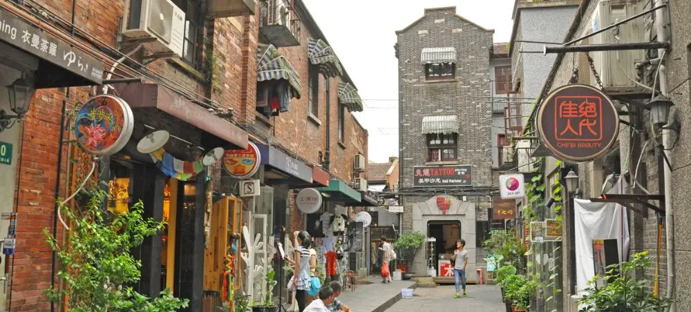 Narrow cobblestone alley in Shanghai's historic district with traditional shops, colorful signage, and visitors exploring local culture