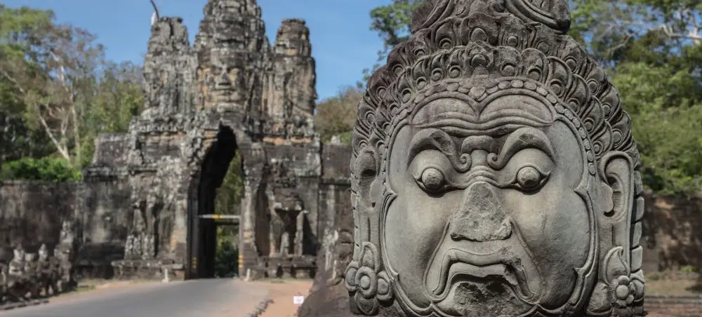 Ancient stone guardian statue at Angkor Thom entrance with Bayon temple towers in Siem Reap, Cambodia